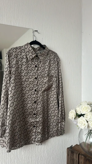 Beige mönstrad skjorta med kedjor - Snygg beige skjorta med coolt kedjemönster i svart. Skjortan har klassisk krage, lång ärm och knappar framtill. Materialet är mjukt och har en lätt glansig finish som ger en lyxig känsla. Perfekt för dig som gillar statement-plagg.