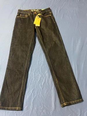 Mörkblå jeansbyxor Rex Regular 152 - Snygga mörkblå jeansbyxor från C Denim, modell Rex Regular i storlek 152. Klassisk femficksdesign med kontrasterande gula sömmar och läderpatch bak. Raka ben och normal passform, perfekta för en clean och stilren look.