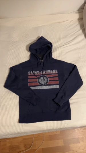 Mörkblå Saint Laurent hoodie - Snygg mörkblå hoodie från Saint Laurent med tryck i vitt och rött på bröstet. Tröjan har huva med snörning och en klassisk känguruficka framtill. Perfekt för dig som vill ha en stilren och exklusiv look med streetvibe. Nyckick och i storlek S
