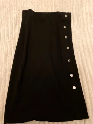 Svart pennkjol med silverknappar - Svart kjol med rak passform och silverfärgade tryckknappar längs sidan. Kjolen har tjock material så det är inte genomskinligt. Kjolens längd är 57 Cm och Bredd bliv 33~cm. Verkligen en efterfrågad plagg som slutade att säljas på New yorker!! Den är stilren som funkar till många olika outfits, en dag och natt plagg! Materialet känns mjukt och stretchigt. 