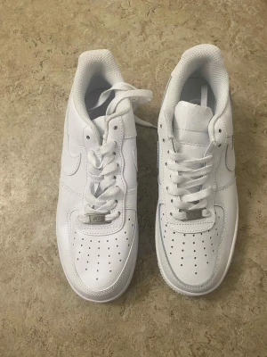 Nike Air Force 1 helvita sneakers - Klassiska Nike Air Force 1 sneakers i helvitt skinn med perforerad tå, rund tå och platt sula. Snygg och clean look med vita snören och diskret Nike-logga på sidan. Perfekta för dig som vill ha en tidlös och fräsch stil.