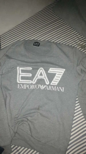 Grå sweatshirt EA7 Emporio Armani - Säljer en grå sweatshirt från EA7 Emporio Armani med stor vit logga framtill. Tröjan har rund hals och långa ärmar, perfekt för en chill och stilren look. Materialet är mjukt och skönt, troligtvis bomull eller polyester. Passar dig som gillar klassiska märken. Broderad text 