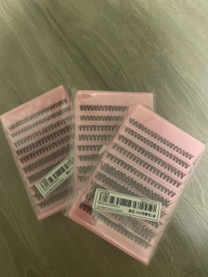 Individuella lösögonfransar i svart - Tre askar med individuella lösögonfransar i svart, förpackade i rosa plastförpackningar. Fransarna är separata och kan appliceras för att skapa en personlig look, perfekt för att bygga volym och längd. Materialet är syntetiskt och varje ask innehåller flera rader med fransar.