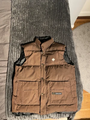 Brun dunväst från Canada Goose - Säljer en brun dunväst från Canada Goose med hög krage, stora fickor framtill och både dragkedja och tryckknappar. Västen har ett stilrent, quiltat utseende och en liten logotyp med lönnlöv på bröstet. Materialet är slitstark polyester med mjukt foder.