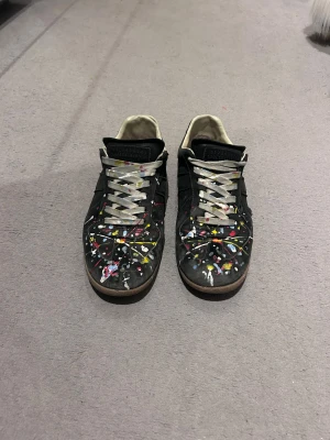  Maison Margiela gats sneakers - Maison Margiela sneakers i svart,  med coolt färgstänksmönster i gult, blått, rött och vitt. Skorna har grå snörning och brun sula. Låg modell med rund tå och unik street-art vibe.  Kan ej bekräfta äktighet, därav priset. Perfekt för dig som gillar statement pieces. Priset kan diskuteras! Hör av er vid frågor eller funderingar! Box tillkommer!