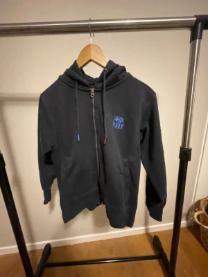 Svart FC Barcelona hoodie XS - Svart hoodie från FC Barcelona i storlek XS. Tröjan har dragkedja framtill, huva med snörning och klubbens logga tryckt på bröstet. Perfekt för dig som älskar fotboll och vill ha en chill look. Passar lika bra till jeans som till mjukisbyxor.