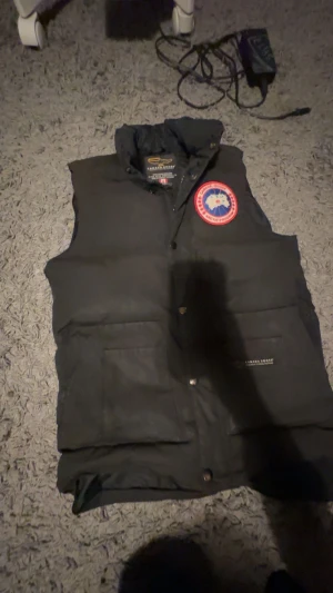 Svart dunväst från Canada Goose - Svart dunväst från Canada Goose med klassisk logga på bröstet. Västen har hög krage, stora fickor framtill och stängs med både dragkedja och knappar. Perfekt för lager-på-lager och riktigt snygg till streetwear-stil.