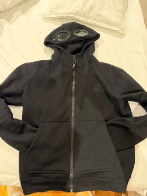 Svart hoodiejacka med goggles CP Company - Svart hoodiejacka från CP Company storlek M men sitter som en S. CP companys ikoniska goggles i huvan. Jackan har dragkedja framtill, två stora fickor och broderad logga på bröstet. Tillverkad i mjuk bomull för skön komfort och streetwear-vibe. Perfekt för dig som gillar unik design och detaljer.