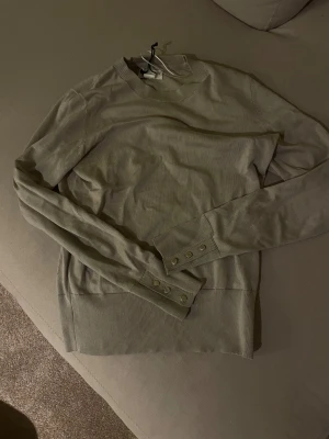 Beige stickad tröja Holly & Whyte S - Beige stickad tröja från Holly & Whyte by Lindex i storlek S. Tröjan har rund halsringning, långa ärmar och snygga knappar vid ärmsluten. Perfekt för lager-på-lager och enkel att matcha med jeans eller kjol.