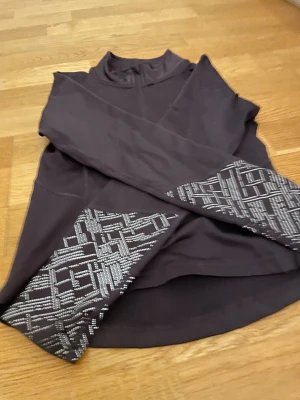 Svart träningströja SOC med mönster - Sportig svart långärmad träningströja från SOC med halv dragkedja och grått grafiskt mönster på ärmarna. Perfekt för träning eller aktiva dagar. Materialet är syntet och designen är smidig och funktionell.