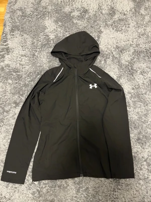 Svart vindjacka från Under Armour - Svart vindjacka från Under Armour med huva och hel dragkedja. Jackan har reflex detaljer på axlarna och logga på bröstet samt stor UNDER ARMOUR-text på ryggen. Tillverkad i lätt polyester, perfekt för träning utomhus. Snygg och sportig look.