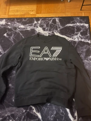 Svart EA7 Emporio Armani sweatshirt - Svart sweatshirt från EA7 Emporio Armani med stor vit logga framtill. Klassisk rund halsringning och långa ärmar. Skön passform och stilren look, perfekt till jeans eller joggers. Materialet känns mjukt och bekvämt mot huden.