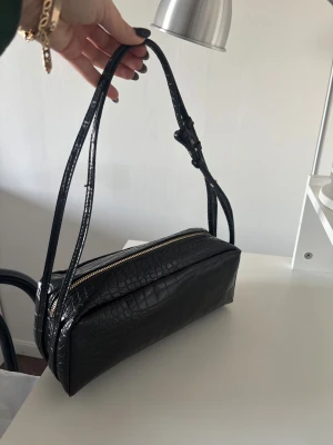 Svart axelväska i croco-imitation - Stilren svart axelväska med croco-mönstrad yta och justerbara smala axelband. Väskan har en rektangulär form och stängs med en guldfärgad dragkedja. Perfekt för dig som gillar en clean och trendig look.