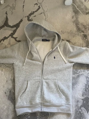 Grå hoodie från Polo Ralph Lauren - Snygg ljusgrå hoodie från Polo Ralph Lauren med dragkedja och huva. Tröjan har två fickor framtill, vit snörning i huvan och den klassiska blå Polo-loggan broderad på bröstet. Mjuk insida i fleece och ribbade muddar vid ärmslut och nederkant.