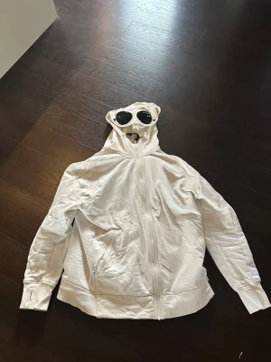 Vit hoodiejacka från C.P. Company  - Vit hoodiejacka från C.P. Company i storlek XL. Jackan har hel dragkedja framtill, två fickor med dragkedja och tumhål i muddarna. Tillverkad i mjuk bomull, perfekt för en chill och clean look.