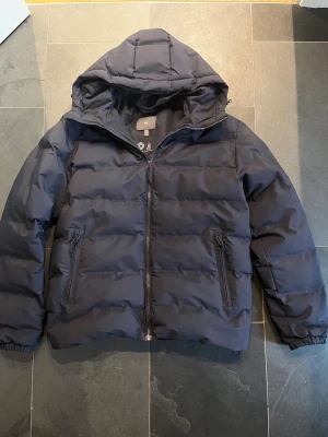 Pufferjacka från HM - Snygg mörkblå pufferjacka från H&M med huva och dragkedja framtill. Jackan är använd ett fåtal gånger förra vintern och har inga defekter. Pris kan diskuteras. Skriv gärna vid funderingar 🤩