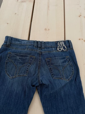 Blå Miss Sixty bootcut jeans - Snygga blå jeans från Miss Sixty med bootcut passform och klassisk femficksdesign. Jeansen har låg midja, slitningar och kontrastsömmar samt en broderad Miss Sixty-logga baktill. Tillverkade i mjukt denim med dragkedja och knapp framtill.