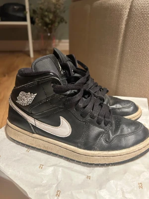 Nike Air Jordan 1 svart/vit strl 38 - Säljer ett par Nike Air Jordan 1 sneakers i svart och vitt med klassisk high-top siluett. Skorna har snörning, rund tå och vit sula med svart yttersula. Ikoniska Air Jordan-loggan på sidan och Jumpman på plösen. Tillverkade i skinn och textil för en snygg streetstil.