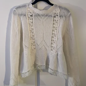 Vit stickad tröja med spetsdetaljer - Supersöt vit stickad tröja med genombrutna mönster och breda spetsdetaljer längs fram och nederkant. Tröjan har långärmade ärmar och en feminin, lätt utsvängd passform. Perfekt för dig som gillar romantisk och mjuk stil.