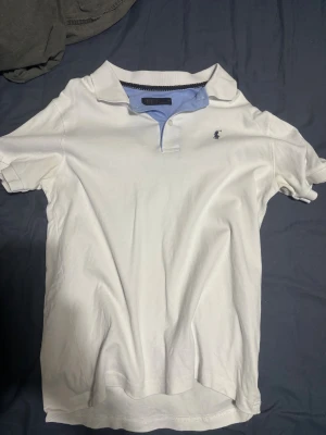 Vit pikétröja från Polo Ralph Lauren - Vit pikétröja från Polo Ralph Lauren med klassisk krage och korta ärmar. Tröjan har en ljusblå insida vid kragen och den ikoniska broderade polospelaren på bröstet. Tillverkad i mjuk bomull och har knappar vid halsen för en clean look. Använd en ända gång!