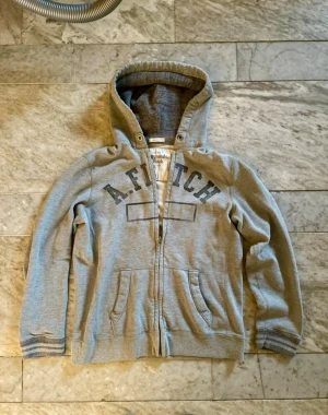 Grå hoodie Abercrombie & Fitch S - Snygg grå hoodie från Abercrombie & Fitch i storlek S. Tröjan har dragkedja, huva med mjukt foder och två fickor framtill. Tryck med A. Fitch över bröstet och ribbade muddar vid ärmslut och nederkant. Perfekt för chill dagar.