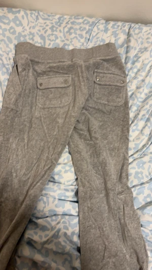 Grå mjukisbyxor från Juicy Couture - Snygga grå mjukisbyxor från Juicy Couture i storlek M. Byxorna har bred resår i midjan, fickor med lock och knapp bak samt sidofickor fram. Mjukt material som är perfekt för chill dagar. Diskret Juicy Couture-logga broderad på ena benet.