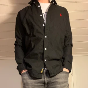 Svart skjorta från Ralph Lauren - Svart skjorta från Ralph Lauren med klassisk krage och vita knappar. På bröstet finns den ikoniska röda broderade Polo-loggan. Perfekt för en clean och stilren look.  Tveka inte på att skriva ifall du undrar nåt!✨