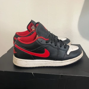 Nike Air Jordan 1 Low Svart/Röd/Vit - Säljer ett par Nike Air Jordan 1 Low med svart läder, röda detaljer och vit tåbox. Skorna har klassisk röd swoosh, svart snörning och röd insida. Ikonisk Jumpman-logga på plösen och Air Jordan Wings på hälen. Perfekta för dig som vill sticka ut med en tidlös sneaker.