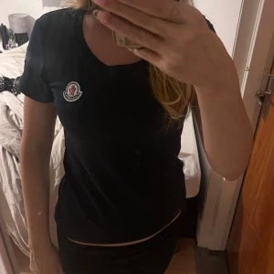 Svart Moncler t-shirt med logga - Snygg svart t-shirt från Moncler med klassisk logga på bröstet och ärmen. Modellen är figurnära med korta ärmar och rund halsringning. ej säker på om det är äkta💗 aldrig använd!