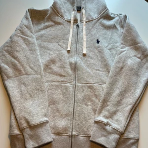 Grå hoodie från Polo Ralph Lauren - Snygg grå hoodie från Polo Ralph Lauren med klassisk broderad logga på bröstet. Tröjan har huva med vita snören, hel dragkedja och ribbade muddar. Perfekt för chill dagar och enkel att matcha med jeans eller joggers.