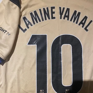 Beige FC Barcelona Lamine Yamal tröja - Säljer en beige FC Barcelona fotbollströja med Lamine Yamal och nummer 10 på ryggen. Tröjan har svarta detaljer, Dri-FIT-material, svarta tryck med Spotify och UNHCR-loggor samt klubbmärke på bröstet. Rund hals med svart och blå kant. Perfekt för dig som älskar Barca.