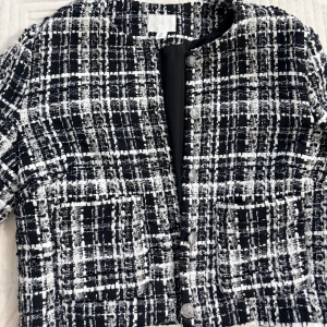 Svartvit rutig kavaj från H&M - Snygg svartvit kavaj från H&M i storlek S med rutigt mönster och silvriga knappar framtill. Jackan har en croppad passform och är tillverkad i ett tjockare, strukturerat tyg som ger en cool och trendig look. Perfekt att styla med jeans eller kjol.