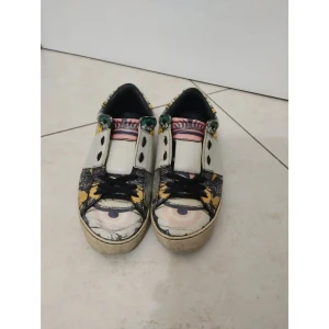 Valentino Garavani sneakers med mönster - Unika sneakers från Valentino Garavani med färgstarkt blommigt och grafiskt mönster i svart, vit, gul och grönt. Skorna har snörning, platt sula och coola nitar runt hälen. Materialet är skinn och detaljerna är riktigt iögonfallande, med ögonmotiv på tån och mönstrad häl. Strl 39