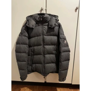Mörkgrå Moncler pufferjacka med huva - Säljer en mörkgrå pufferjacka från Moncler med huva och snygga detaljer som dragkedja och tryckknappar vid ärmslut. Jackan har Moncler-logga på ärmen och två praktiska fickor med dragkedja. Perfekt för dig som vill ha en stilren och varm jacka. Storlek 5 vilket motsvarar XL
