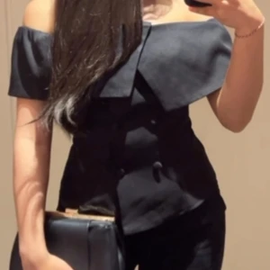 Svart offshoulder korsettopp - Snygg svart korsettopp med offshoulder-design och bred krage som ger en cool och elegant look. Toppen har figurnära passform och knappar framtill för extra detalj. Perfekt för dig som vill sticka ut med en stilren och trendig topp.