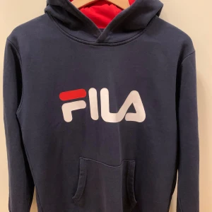 Mörkblå Fila hoodie med röd huva, stl 158/164 - Snygg mörkblå hoodie från Fila med stor logga i vitt och rött på bröstet. Insidan av huvan är röd vilket ger en cool kontrast. Tröjan har känguruficka och långa ärmar med muddar. Perfekt för dig som gillar sportig och avslappnad stil.