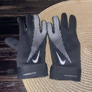 Nike Therma-Fit svarta träningshandskar - Svarta Nike Therma-Fit handskar med vitt mönster och logga på ovansidan. Handskarna har ett sportigt snitt, greppytor på insidan och är tillverkade i syntetmaterial som håller värmen. Perfekta för träning utomhus under kyliga dagar.