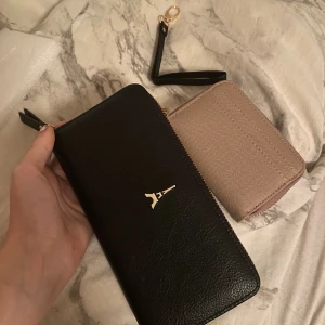 Svart och beige plånbok med dragkedja - Två snygga plånböcker, en i svart skinn med guldfärgad Eiffel-torn detalj och dragkedja, samt en mindre beige plånbok i strukturerad syntet med guldfärgad dragkedja och kortfack. Perfekt för att hålla koll på kort och cash. 2 oanvända plånböcker som inte har märken eller något o man kan köpa separat för 150 kr styck 