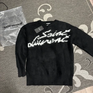 Svart stickad Saint Laurent-tröja S - Exklusiv svart stickad tröja från Saint Laurent Paris med vit logotyp i graffiti-stil över bröstet. Tröjan har rund halsringning, långa ärmar och ribbade muddar. Tillverkad i Italien och känns riktigt lyxig och mjuk.