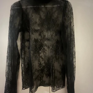 Svart spetsig polotopp från Soyaconcept - Säljer en svart, transparent polotopp från Soyaconcept i storlek S. Toppen har lång ärm, hög krage med volangkant och är helt täckt av ett snyggt spetsmönster. Perfekt för att ge din outfit en cool och edgy vibe.