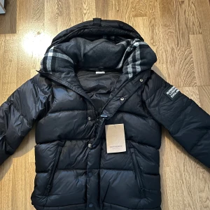 Burberry Puffer Jacka - Säljer en svart pufferjacka från Burberry med avtagbara ärmar. Jackan har dragkedja, tryckknappar och flera fickor. Insidan av huvan har klassiskt rutigt Burberry-mönster. Perfekt för kalla dagar och riktigt snygg streetstil.