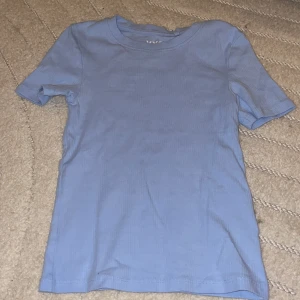 Ljusblå ribbad t-shirt FSBN Sister - En basic ljusblå t-shirt från FSBN Sister i ribbad bomull. Modellen är kortärmad med rund halsringning och har en clean, enkel look utan tryck eller mönster. Perfekt för dig som gillar stilrena plagg i mjukt material.