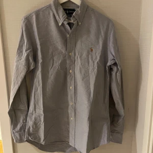 Grå skjorta från Ralph Lauren - Snygg grå skjorta från Ralph Lauren med klassisk button-down krage och broderad logga på bröstet. Skjortan har lång ärm, vita knappar och är i bomull med en custom fit-passform. Perfekt för dig som gillar stilrena och tidlösa plagg. Sitter som M