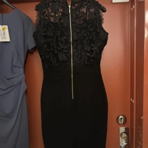 Svart spetsklänning med dragkedja - Snygg svart kortklänning med spets upptill och figurnära passform. Klänningen är ärmlös och har en dekorativ dragkedja baktilll,. Spetsen har ett blommigt mönster som ger en elegant touch. Perfekt för dig som vill ha en stilren look med lite edge.