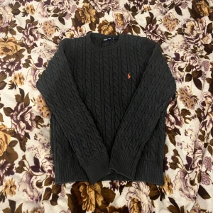 Mörkgrå stickad tröja från Ralph Lauren - Snygg mörkgrå kabelstickad tröja från Ralph Lauren i storlek M. Tröjan har rund hals, långa ärmar och klassisk orange broderad logga på bröstet. Perfekt för dig som gillar stilrena och tidlösa plagg med premiumkänsla. Storlek M men passform S / XS