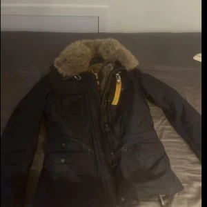 Svart parkas från Parajumpers med päls - Säljer en mörkblå jacka från Parajumpers med fluffig brun päls runt kragen och insidan. Jackan har flera fickor, gul detalj vid dragkedjan och patch på ärmen. Perfekt för kalla dagar med sin quiltade insida och snygga look.