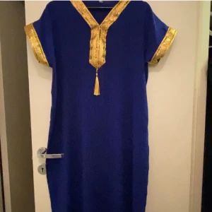 Blå klänning med gulddetaljer - Snygg blå klänning med korta ärmar och guldfärgade detaljer vid halsringning och ärmslut. Klänningen har en rak passform och v-ringad hals med dekorativ tofs framtill. Perfekt för dig som gillar färgstarka och unika plagg.det passar folk L M S 