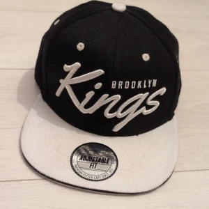 Brooklyn Kings keps från FSBN - Snygg snapback-keps från FSBN med broderad text 'Brooklyn Kings' i vitt. Kepsen är svart med vit skärm och vita detaljer, samt justerbar passform baktill. Extra broderade detaljer på sidan och baksidan med 'Sports Edition'. Perfekt för en sportig streetstyle.