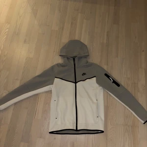 Nike hoodie i grått och vitt fleece - Nike hoodie i mjuk fleece med grått upptill och vitt nedtill. Svarta detaljer längs dragkedjan och ärmslut, samt en praktisk ficka med dragkedja på ena ärmen. Huva och långärmad design, perfekt för chill eller träning. Storlek S men passar lika bra på folk med storlek M