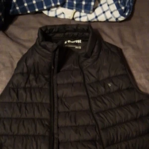 Svart dunväst från Everest - Svart dunväst från Everest i storlek 146/152. Västen har quiltad design med horisontella sömmar och dragkedja framtill. Perfekt för lager-på-lager och har en stilren look som funkar till många outfits.
